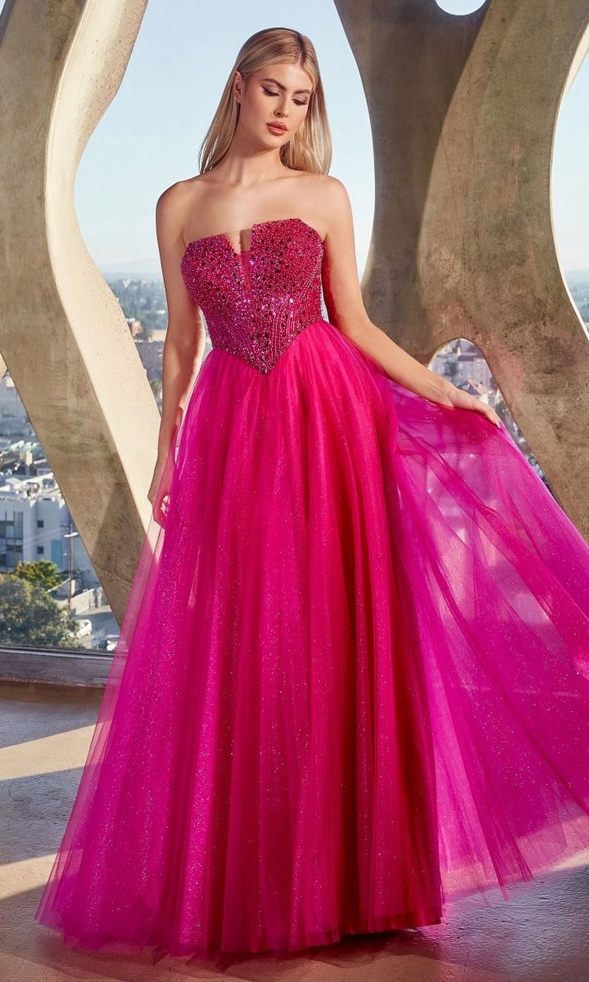 A woman in the Lace-Up Strapless Long Glitter Prom Dress CD0217—a sparkling bright pink, floor-length gown with a beaded bodice and tulle skirt—stands indoors by large windows.